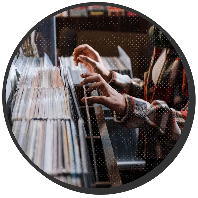 Young person wearing a flannel shirt is flipping through records in a record store. There are headphones dangling around their neck.