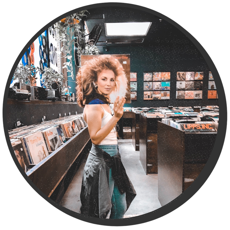 Woman standing in a record store isle, holding a record over her face to personify the artist on the album cover. The woman on the record cover has very cool 80's rocker hair.